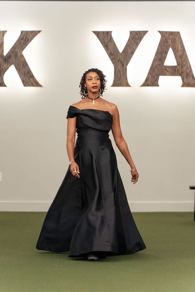 Woman in a black off-the-shoulder gown walks on a green carpeted runway, logo in the background.