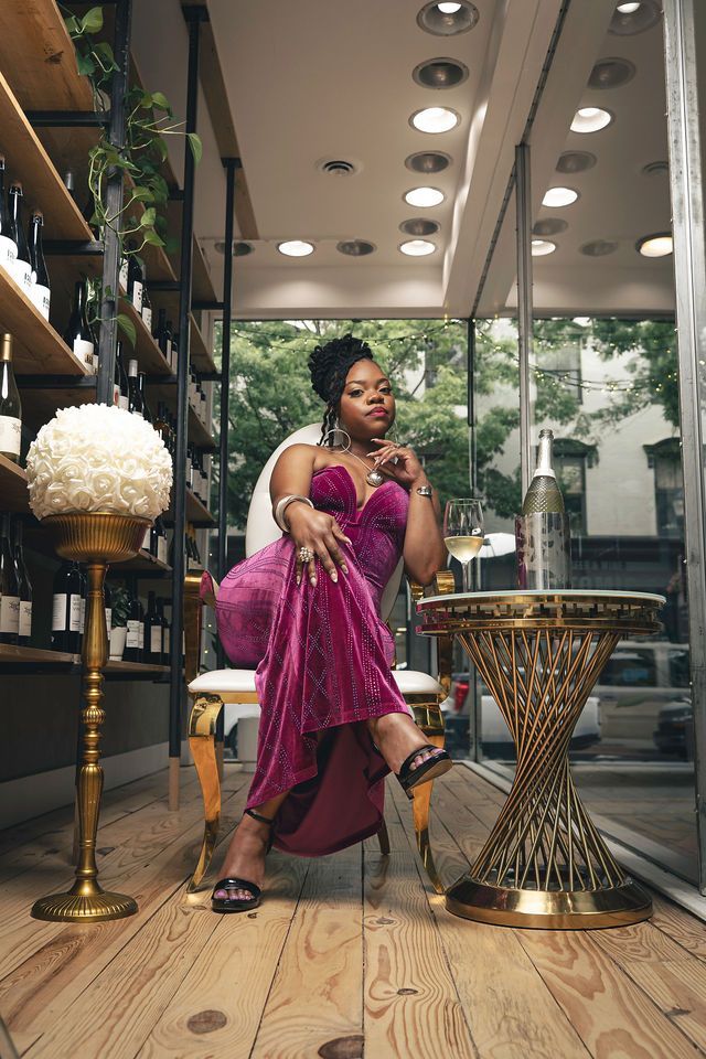 Woman in a pink velvet dress sits in a wine shop, next to a gold table and floral arrangement.