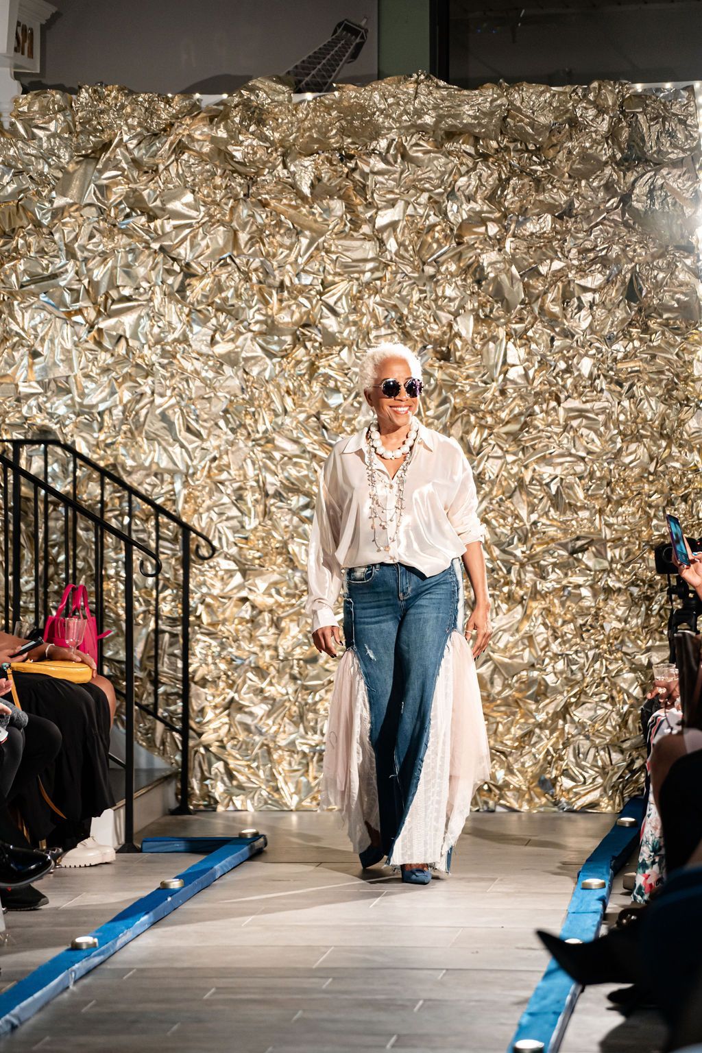 Fashion show model walks runway in front of a golden background; wearing a white top, jeans, and sunglasses.