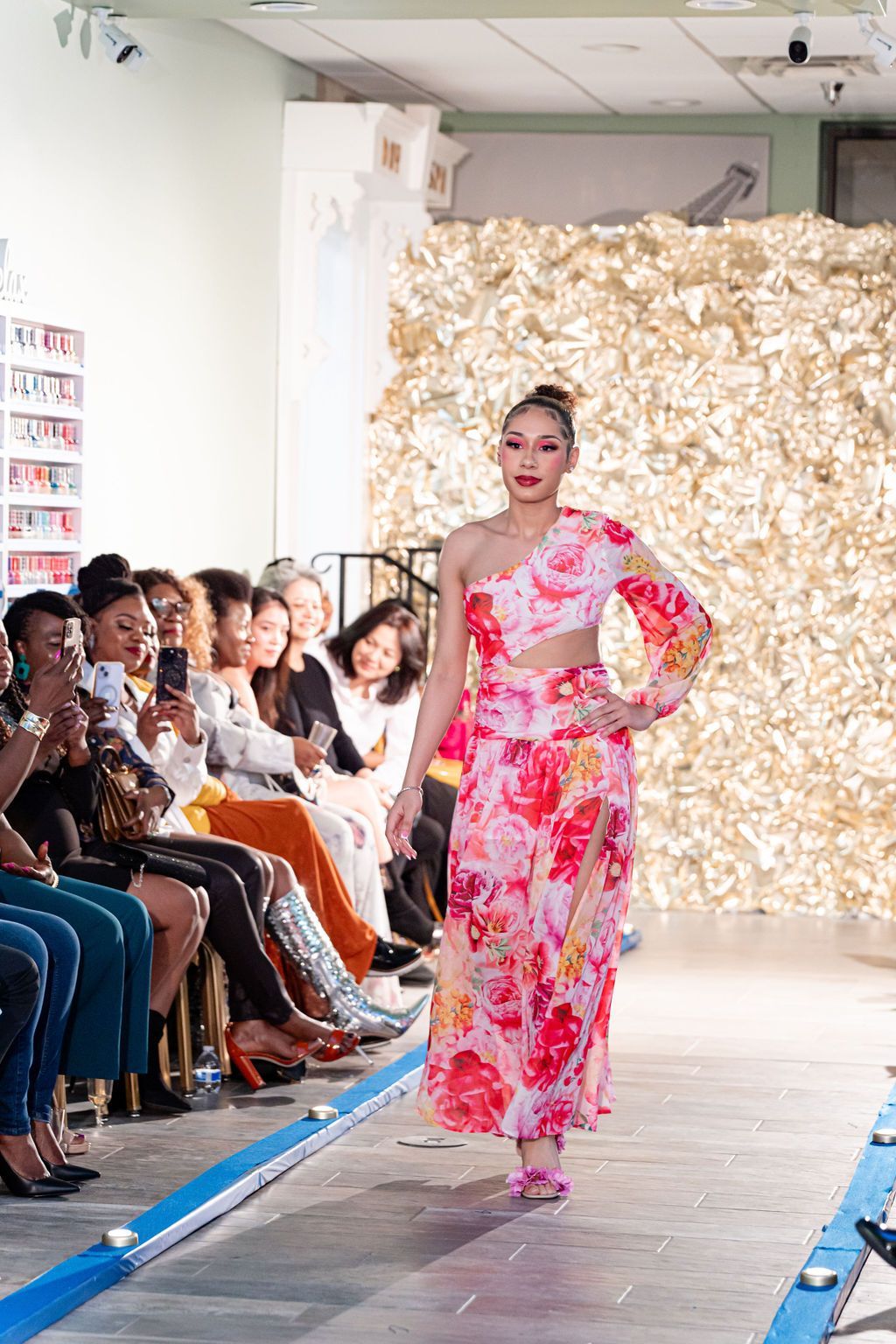 Woman in pink floral dress walks a fashion runway. Audience watches.