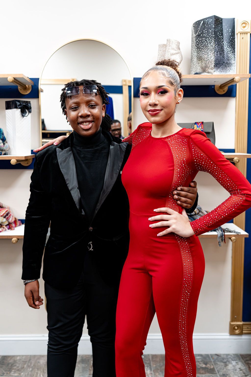Two people pose together in a clothing store. One wears red jumpsuit, other black suit.