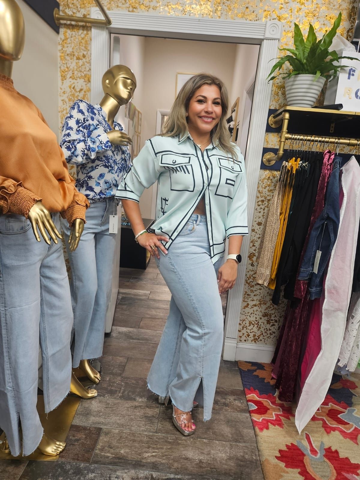 Woman in a boutique, wearing a light blue top and jeans, smiling. Mannequins in the background.