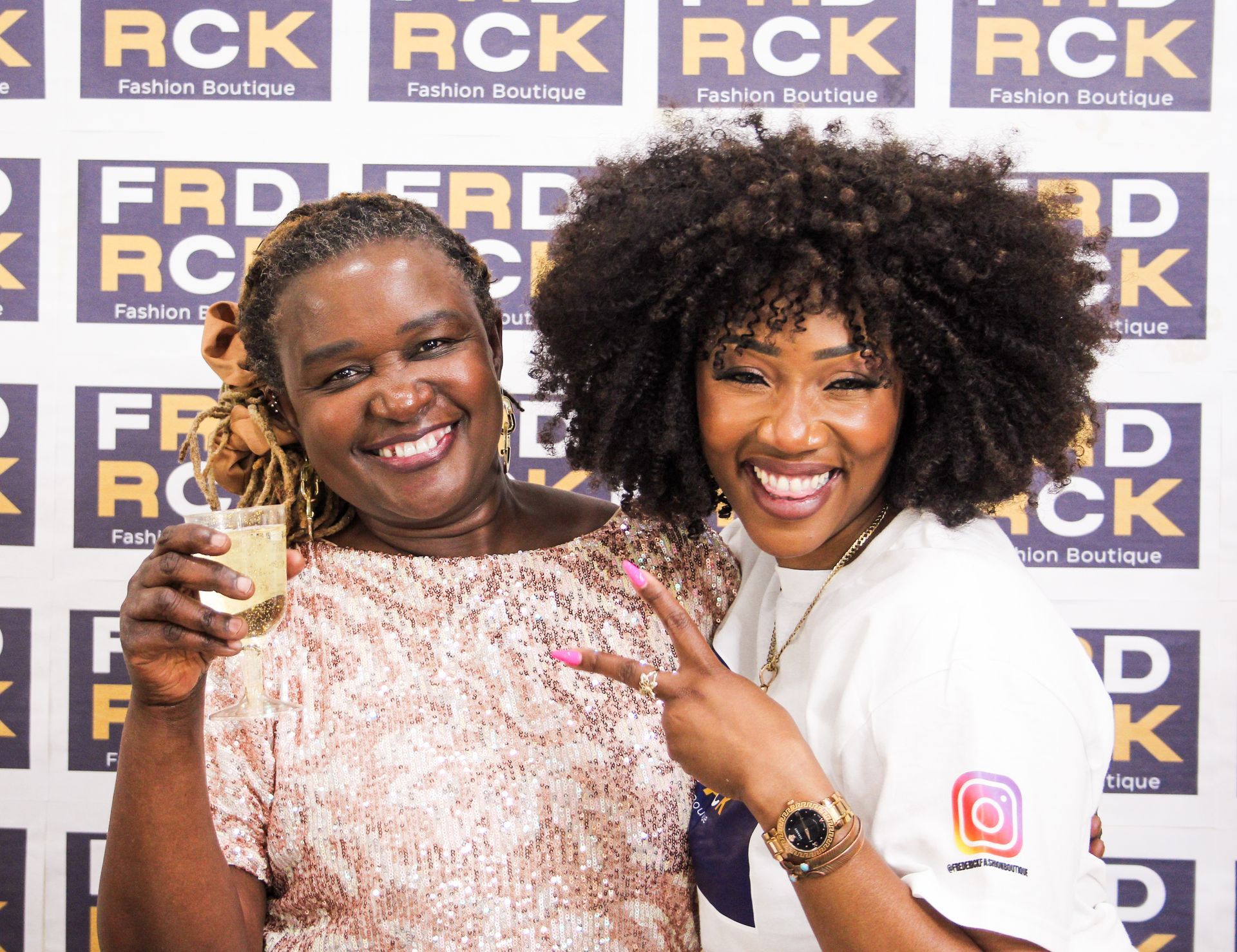 Two smiling women posing in front of a backdrop. One holds a drink, the other makes a peace sign.