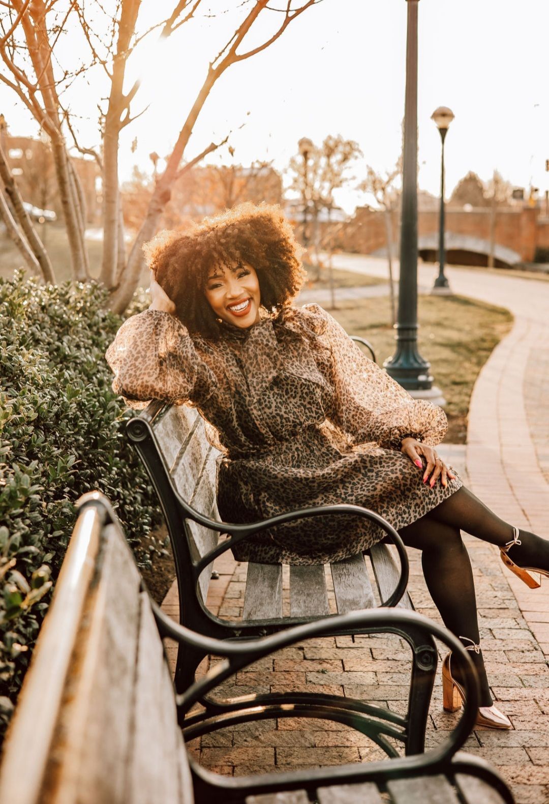 Woman smiling, leaning on park bench, wearing patterned dress, tights, and heels.