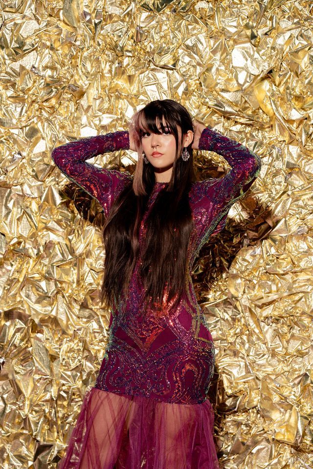 Woman in sequined purple dress with hands on head, against a gold textured background.