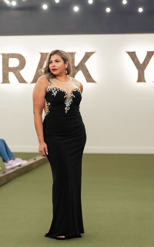 Woman in black evening gown, posing on green surface, under a white wall with letters.