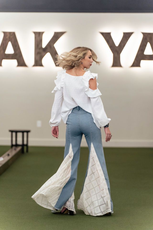 Woman in white top and flared jeans, standing on green turf, hair blowing.