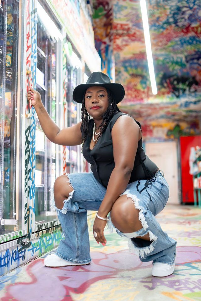 Woman in a black hat, vest, and ripped jeans squats by a graffiti-covered cooler in an art-filled space.