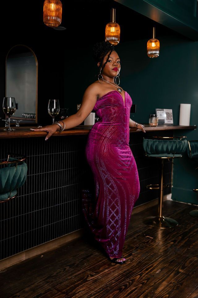 Woman in a magenta strapless gown leans on a dark bar with gold accents.