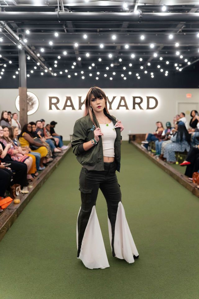 Fashion model in olive green jacket, white top, and pants with white flares on green runway; audience in background.