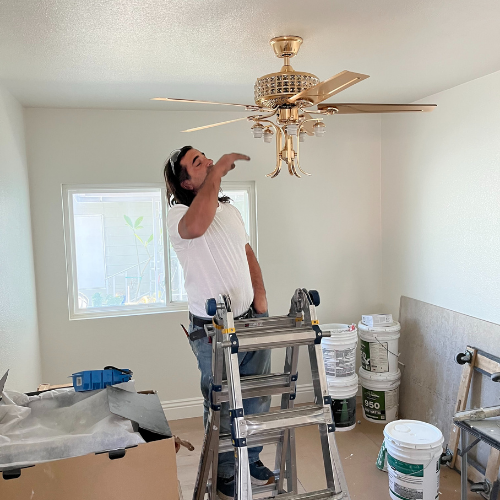 A man is standing on a ladder looking up at a ceiling fan.