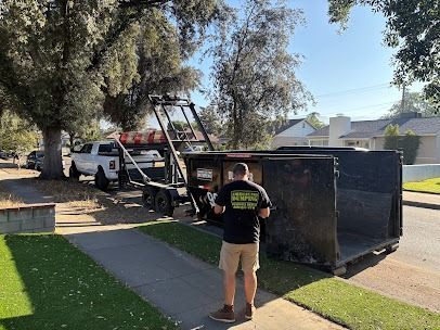 Man standing near an open dumpster on a residential street; a white pickup truck with a container is in the background.