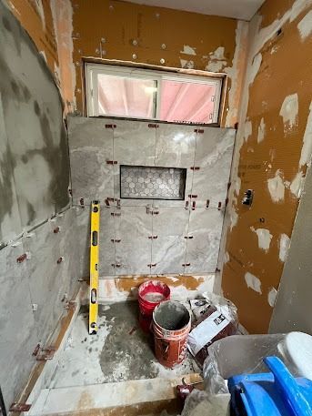 Bathroom under construction; tiles partially installed around a recessed niche.  Orange walls, window, tools, and buckets present.