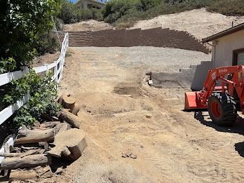 Dirt hillside with retaining wall, white fence, Kubota tractor, and cut logs.