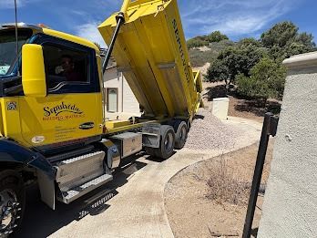Yellow dump truck unloading gravel on a driveway; California home in the background.