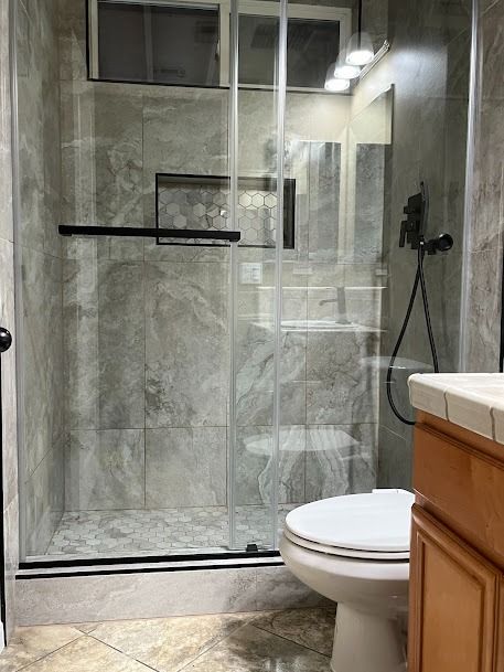Bathroom with a glass shower, toilet, and vanity; grey tile walls, light-colored wood cabinets.