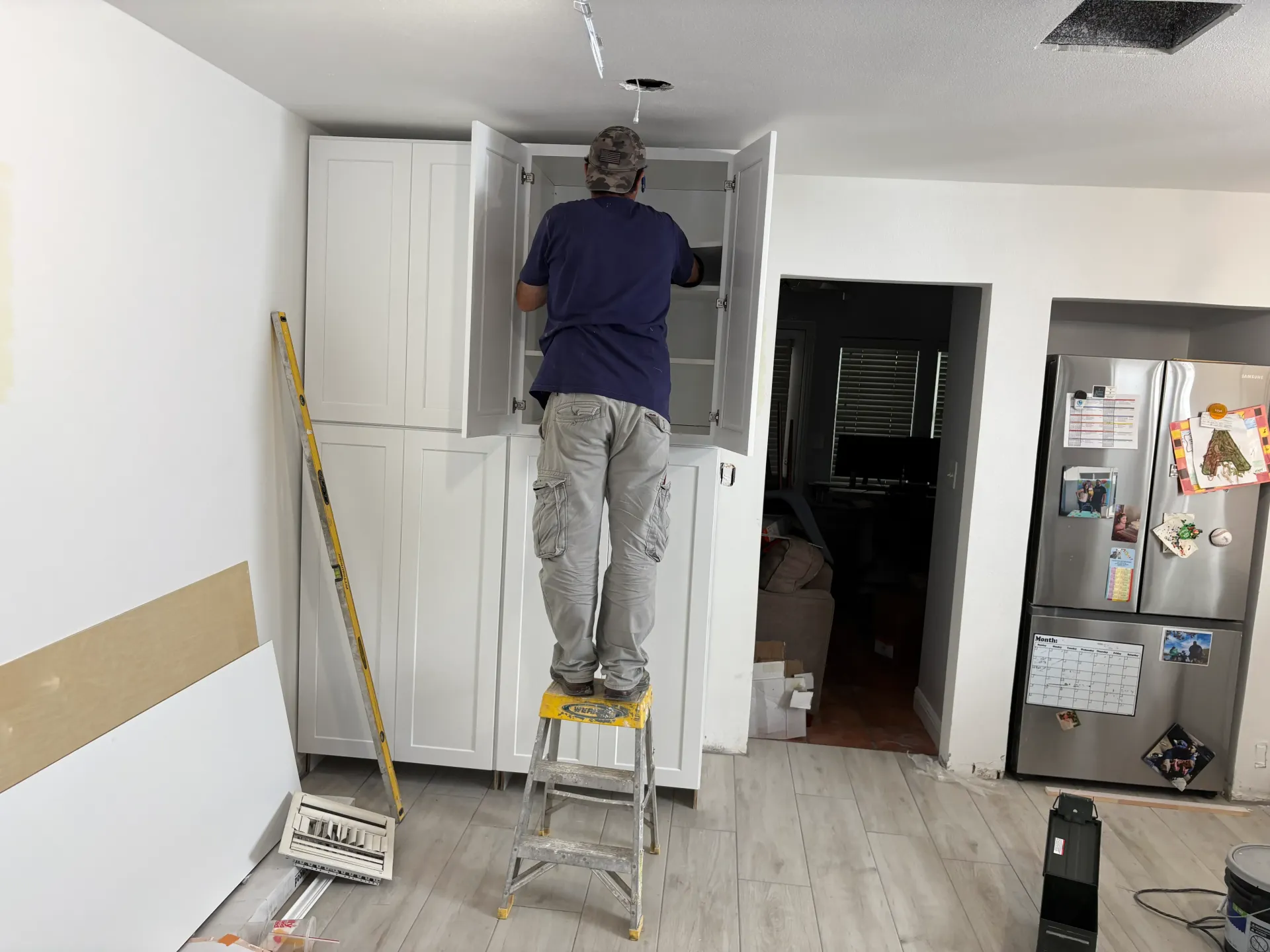 Person on stepladder installing kitchen cabinet in a room with white walls and light flooring.