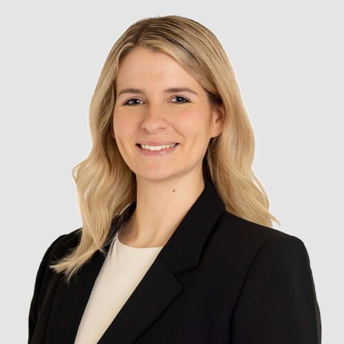 Woman with blonde hair, smiling, wearing a black blazer and white top, against a grey background.