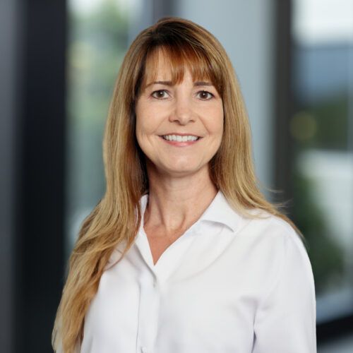 Woman with dark hair smiles, wearing a dark blazer, against a light gray background.