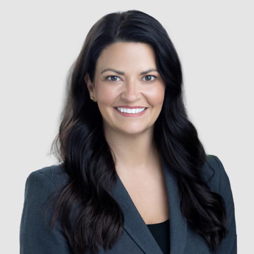 Woman with dark hair smiles, wearing a dark blazer, against a light gray background.