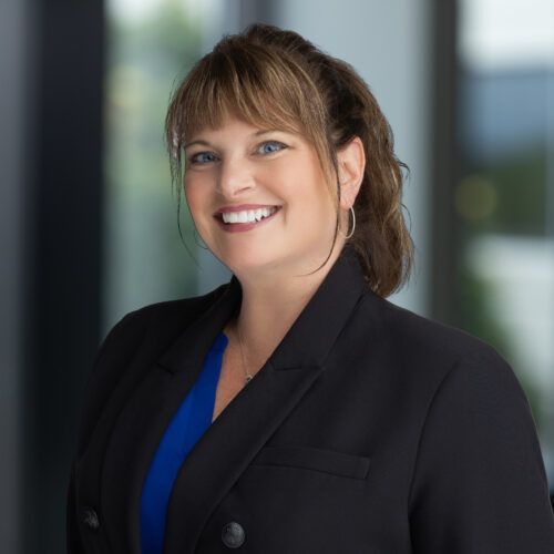 Woman with dark hair smiles, wearing a dark blazer, against a light gray background.
