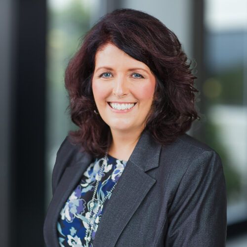 Woman with dark hair smiles, wearing a dark blazer, against a light gray background.