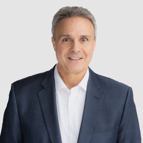 Man in a suit smiles, white shirt, grey hair, studio shot.