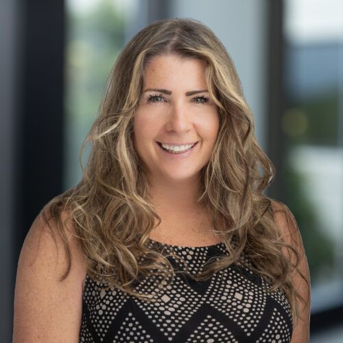 Woman with dark hair smiles, wearing a dark blazer, against a light gray background.