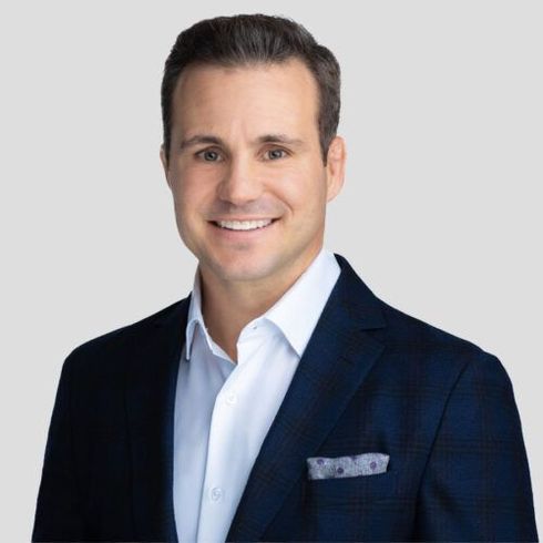 Man in a navy blazer and white shirt, smiling. Gray background, pocket square.