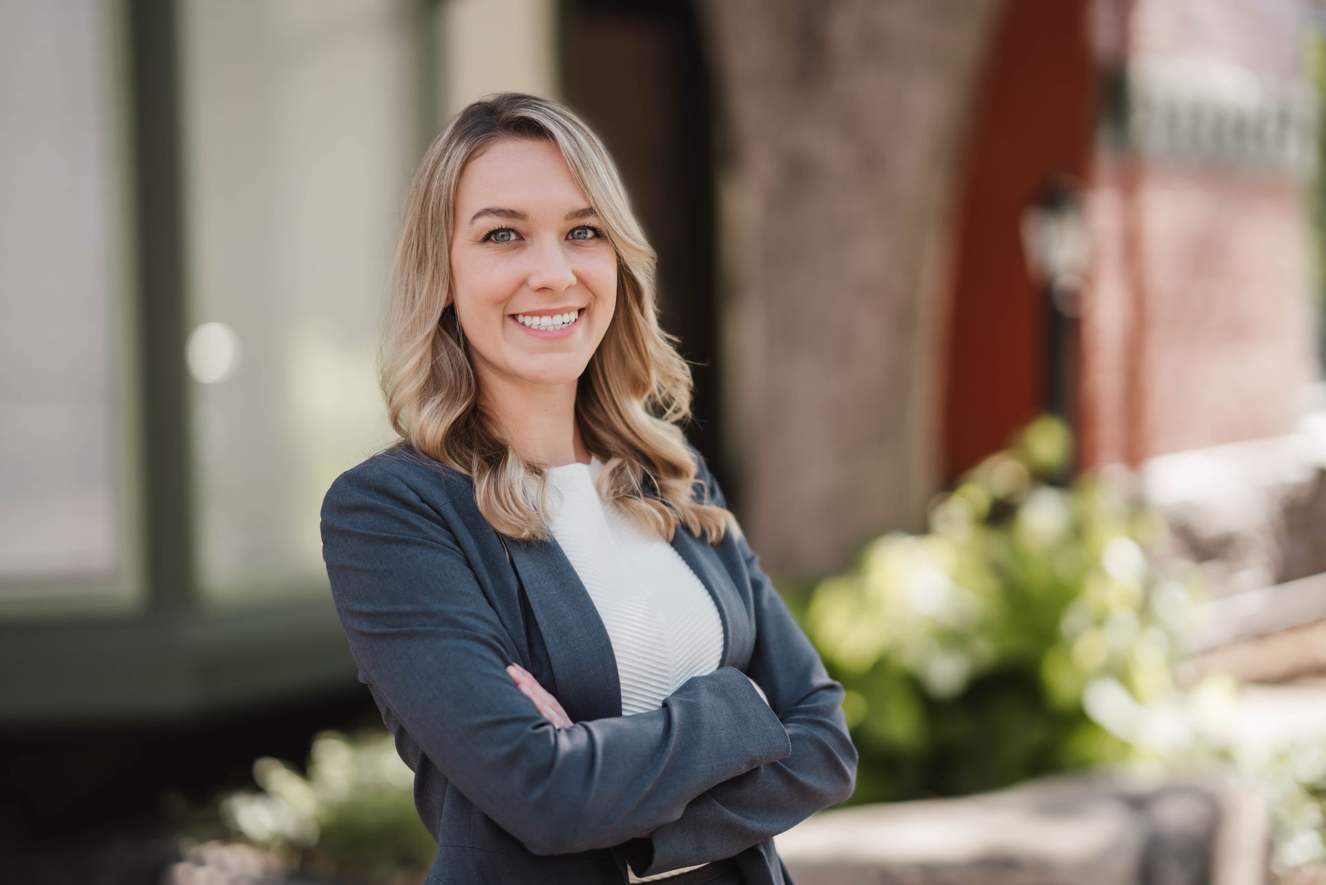 A woman in a suit is standing with her arms crossed and smiling.