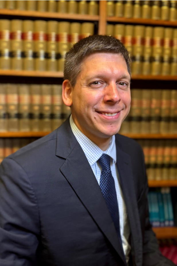 A man in a suit and tie is smiling in front of a bookshelf filled with books.