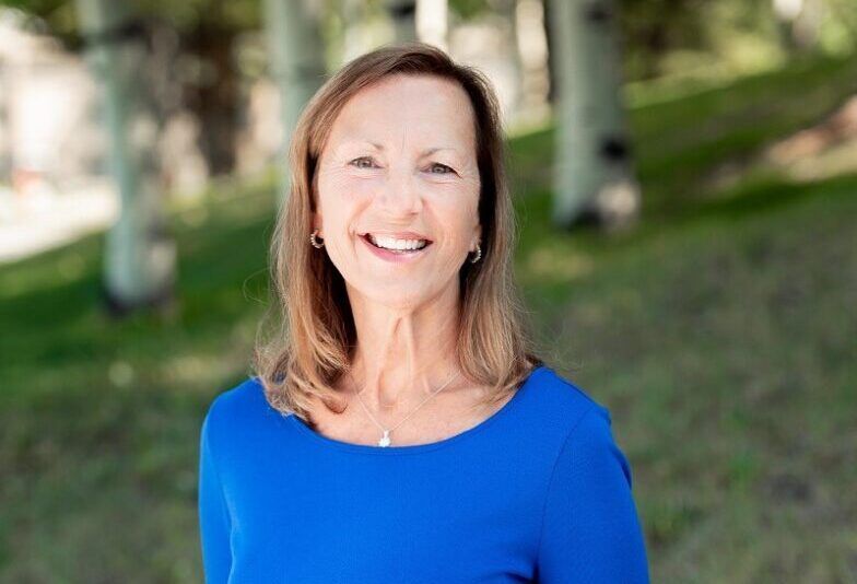 A woman in a blue shirt is smiling for the camera while standing in front of trees.