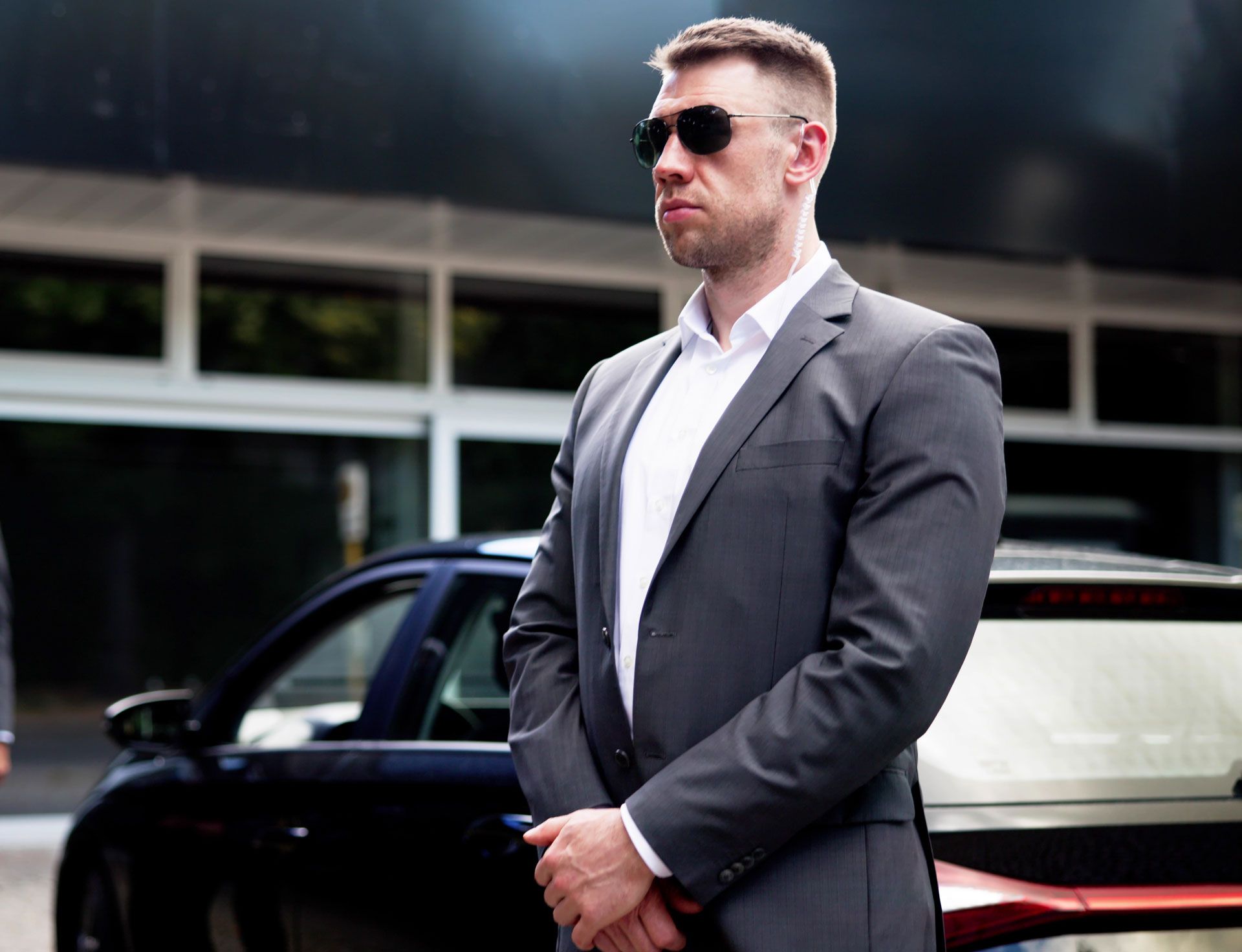 Man in a suit and sunglasses stands in front of a black car, looking vigilant.