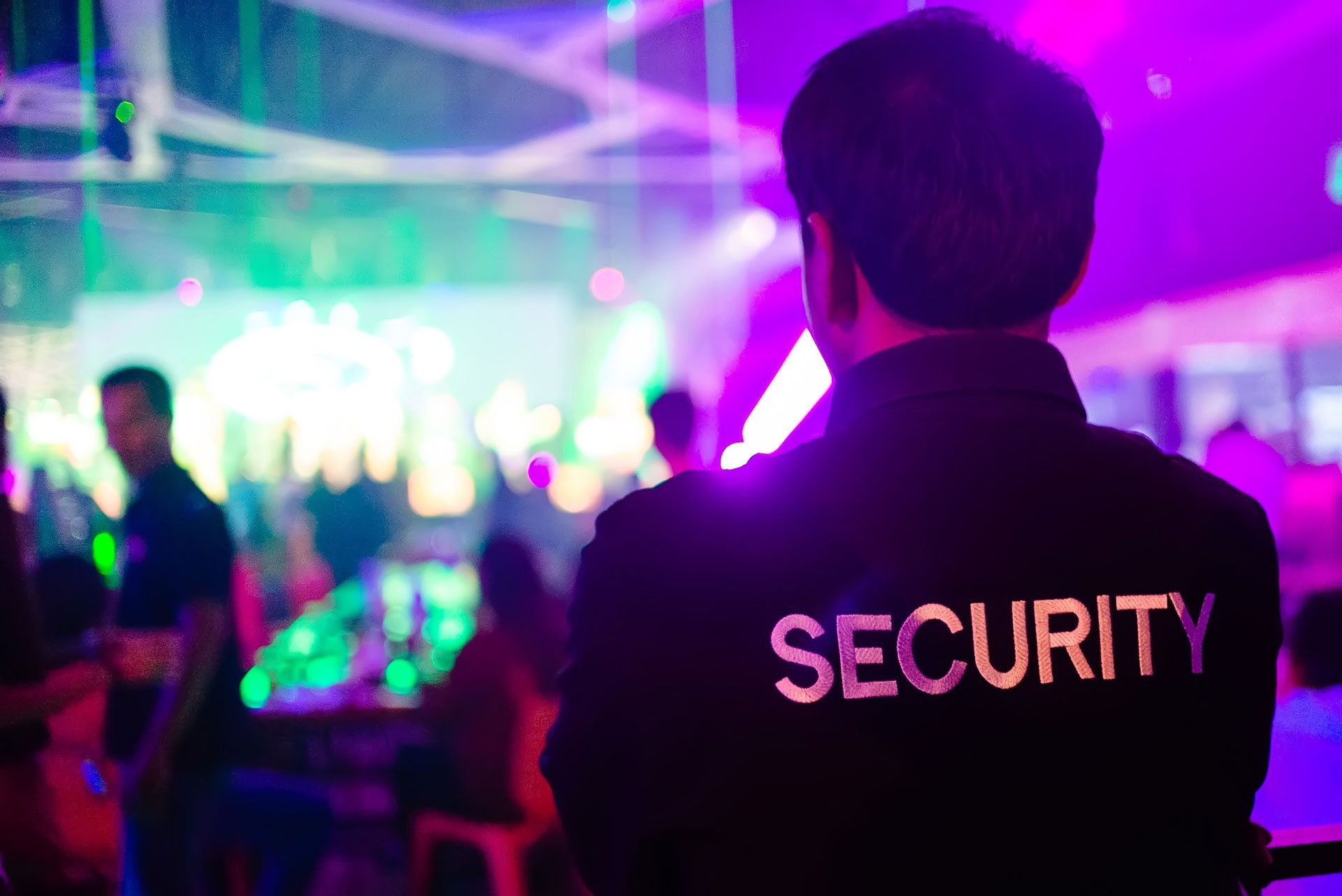 Security guard at a nightclub, back to camera, watching the crowd.  Purple and green lighting.