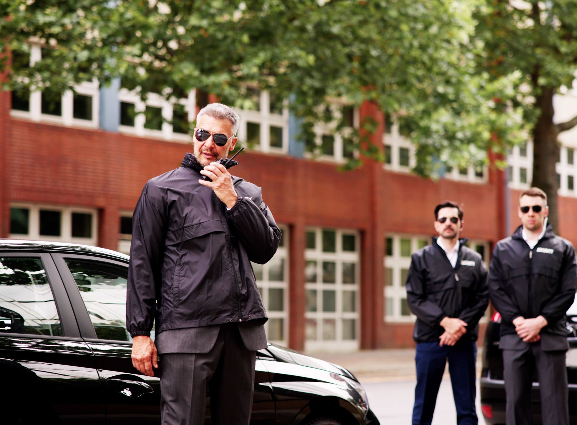 Three security guards near a black car and a brick building; one using a radio.