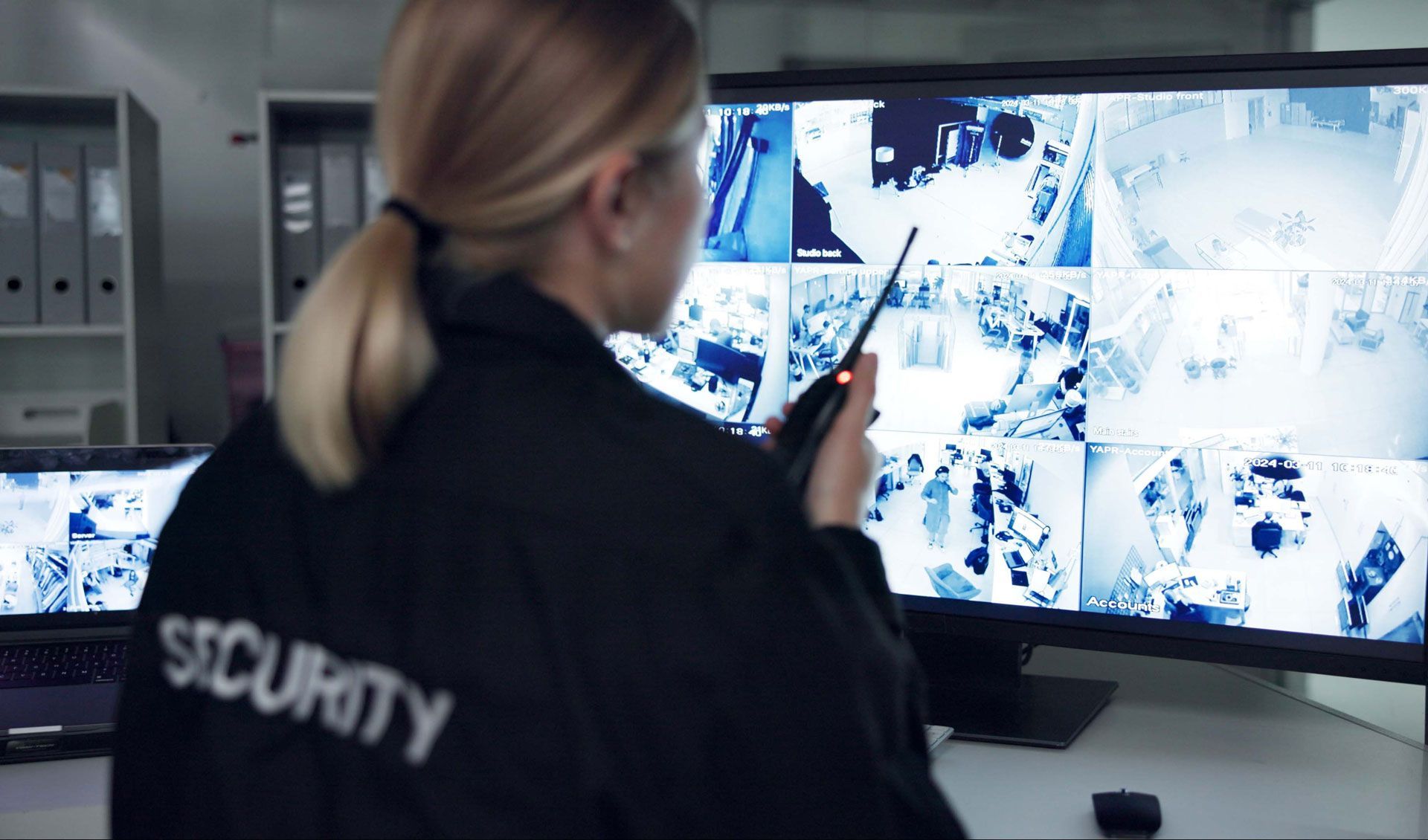 Security guard monitoring surveillance screens, holding a radio, in an office setting.