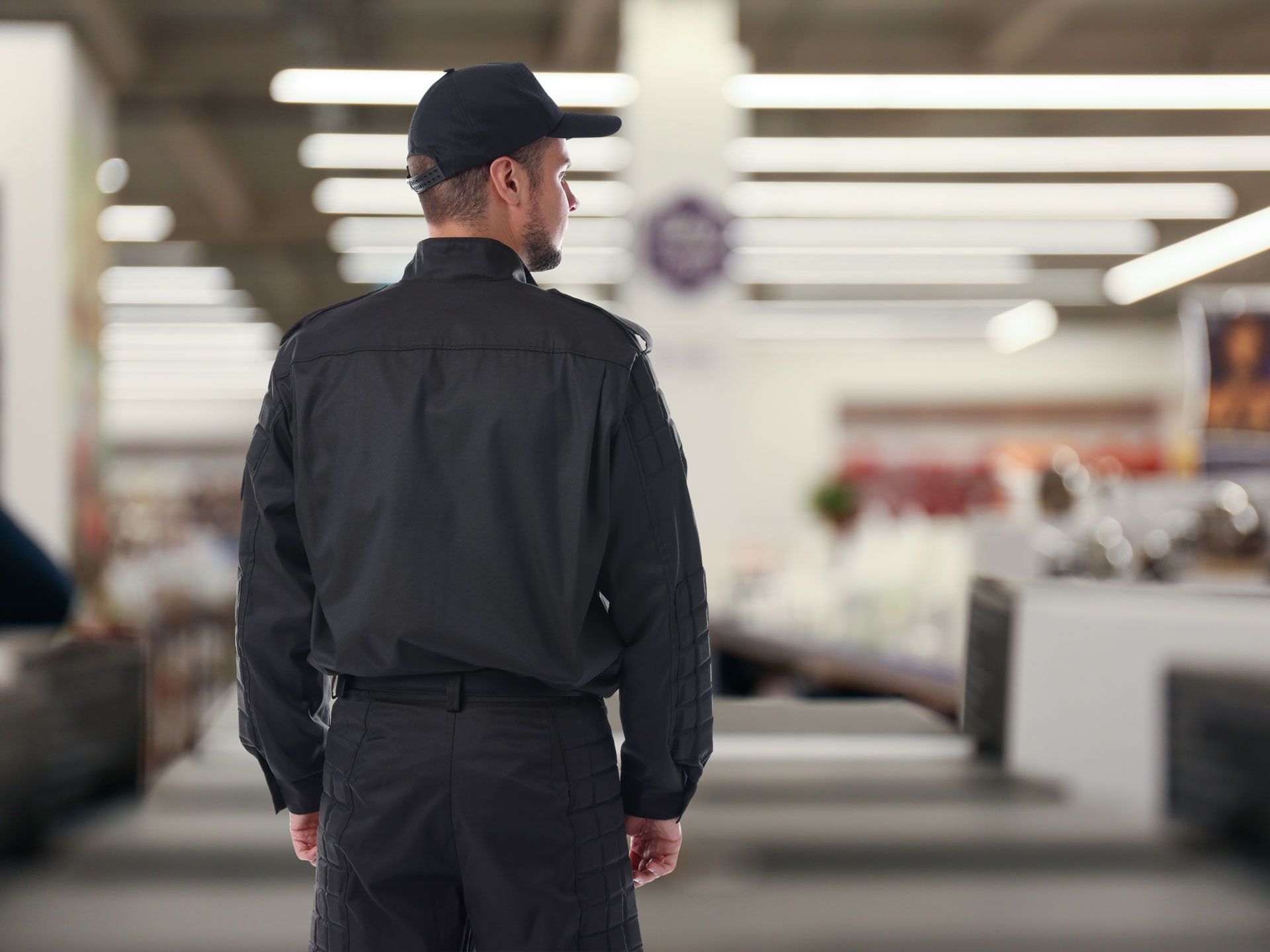Security guard in black uniform and cap, standing in a brightly lit store.