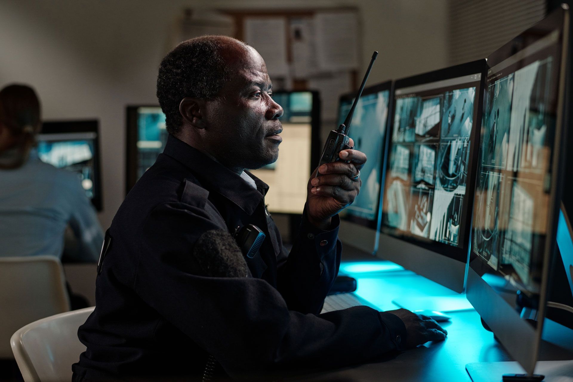 Security guard at computer monitors, holding a radio and looking at screens. Dimly lit room.