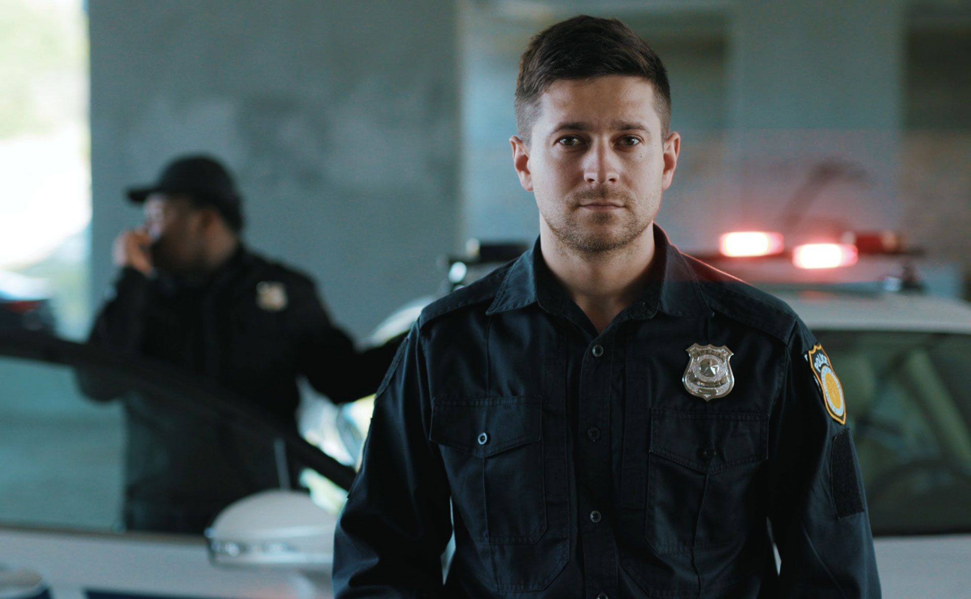 Police officer in uniform stands in front of a police car with lights on; another officer talks on the phone.