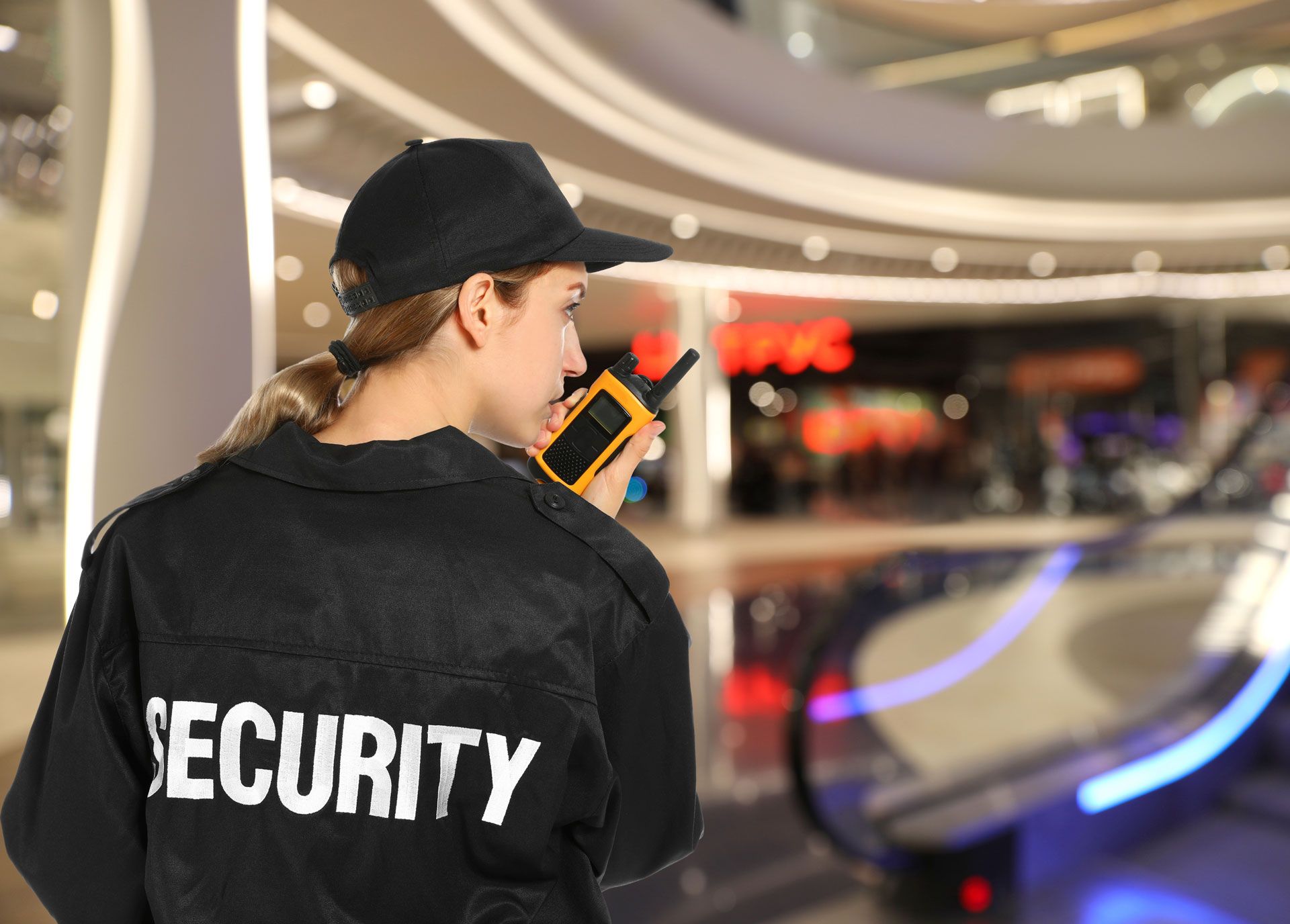 Security guard using a radio in a mall, wearing a black uniform and cap.