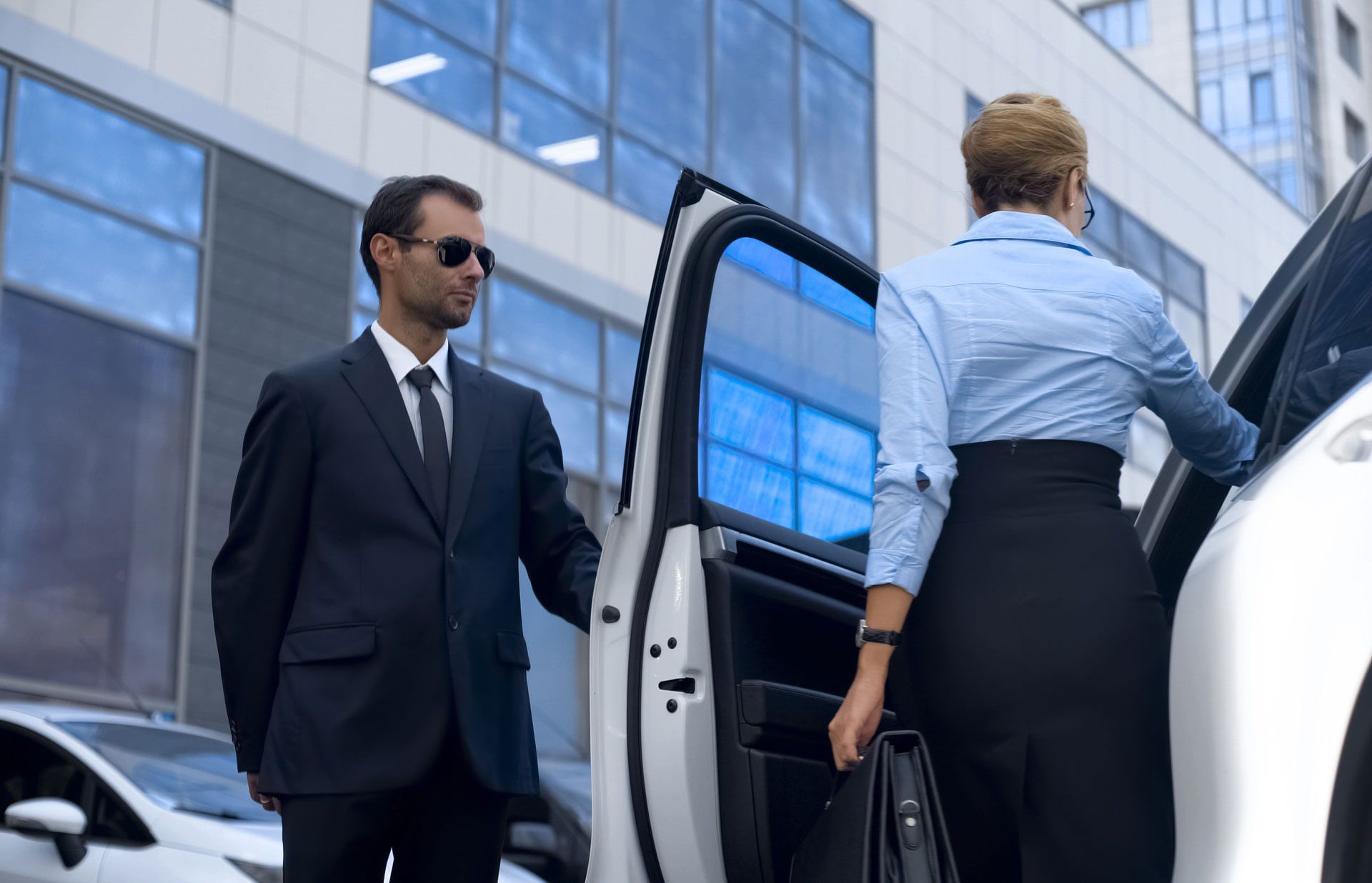 Man in suit opening car door for a person in a skirt, near a building.