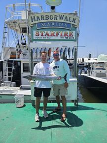 Two men holding a large fish in front of a harbor walk marina sign