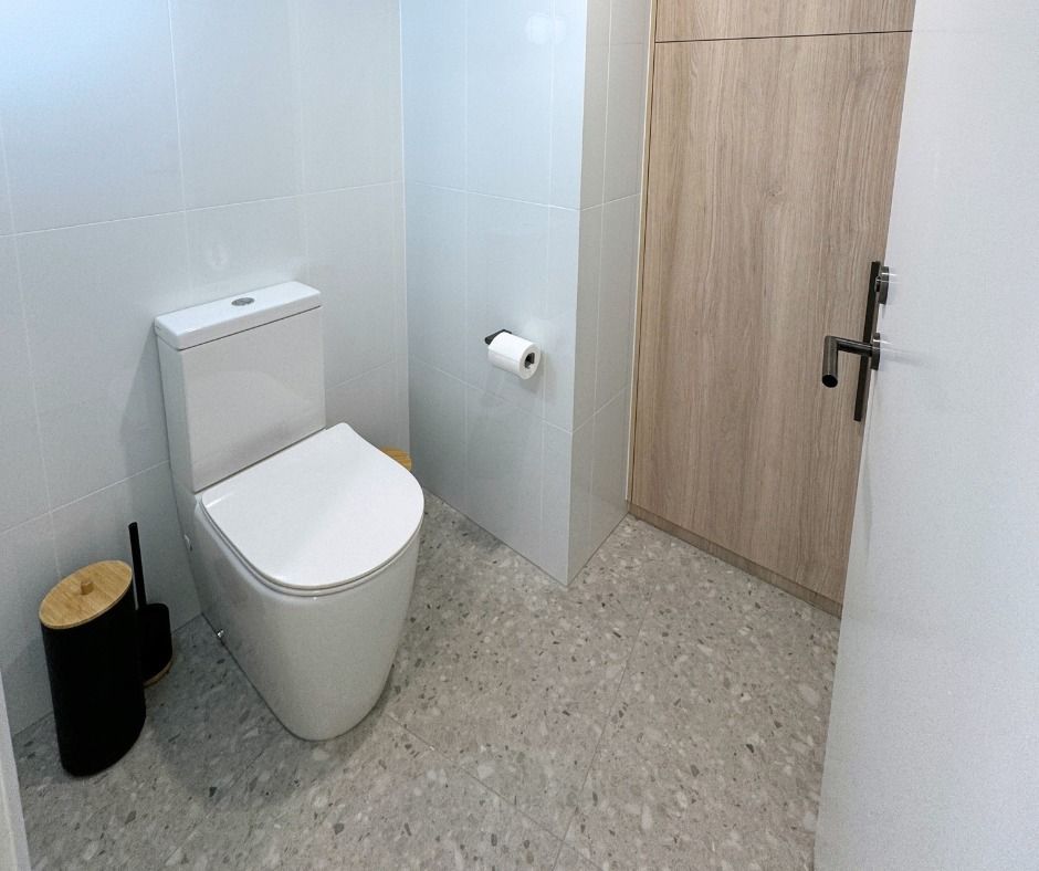 White Toilet in a Small Bathroom With Light Grey Speckled Floor and a Light Wood Door — Nyblad Construction in Buderim, QLD