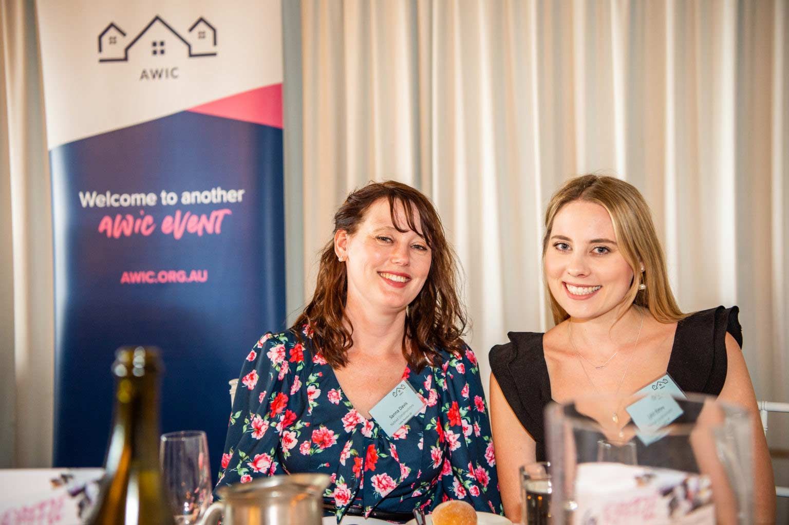 Two Women Are Sitting At A Table With A Sign In The Background — Nyblad Construction in Caloundra West, QLD