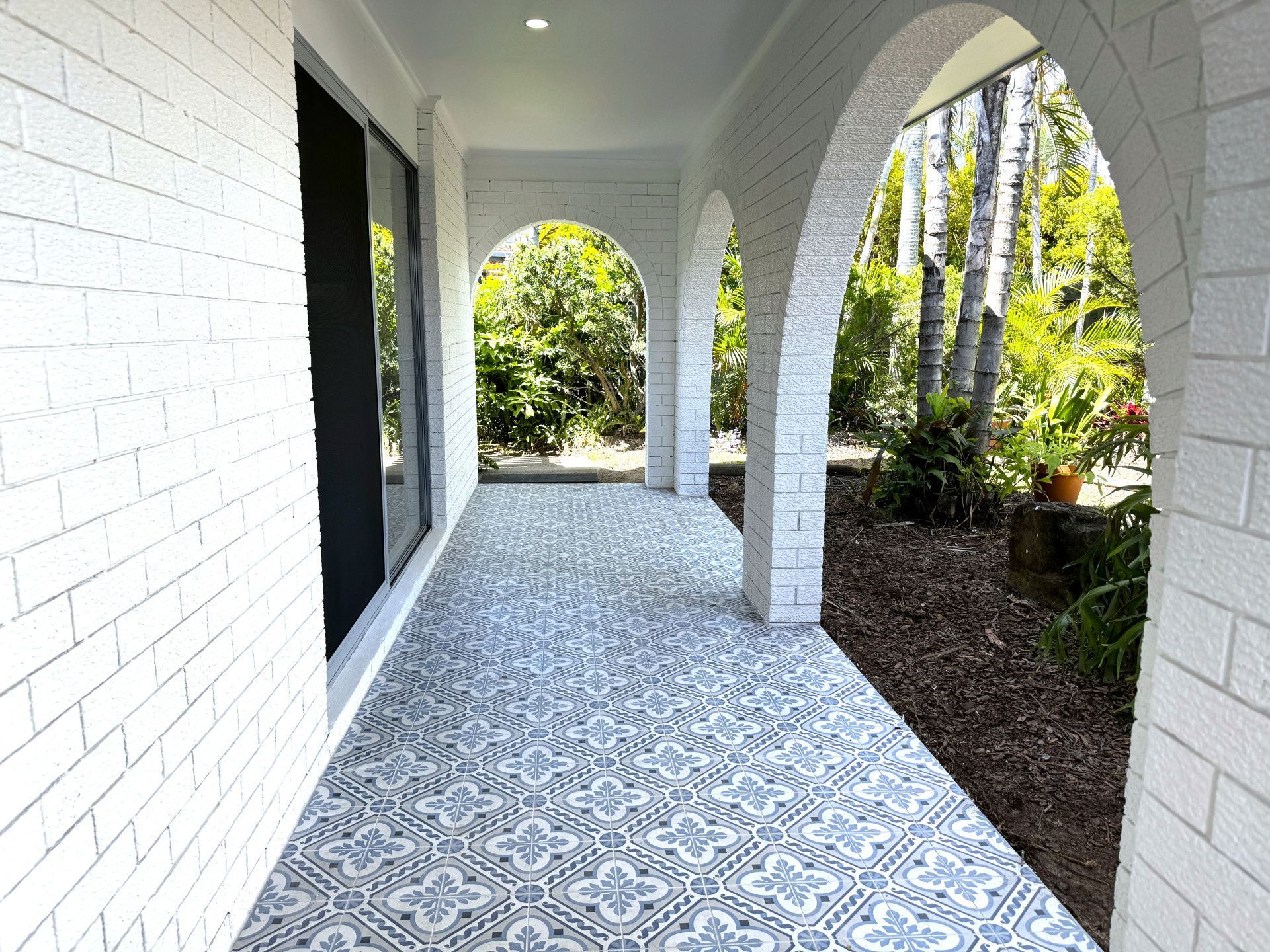 Covered outdoor walkway with white brick walls, arched openings, patterned floor tiles, and greenery. — Nyblad Construction in Coolum, QLD