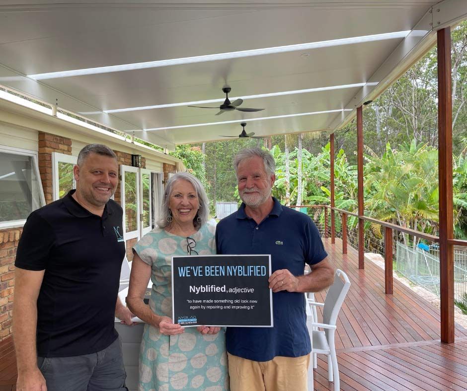 A Man And Two Women Are Standing On A Porch Holding A Sign — Nyblad Construction in Caloundra West, QLD