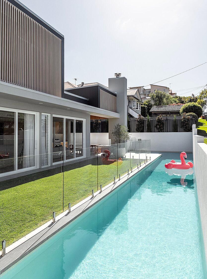Modern backyard with a long turquoise pool beside a house and lawn, with a pink float and white chairs nearby