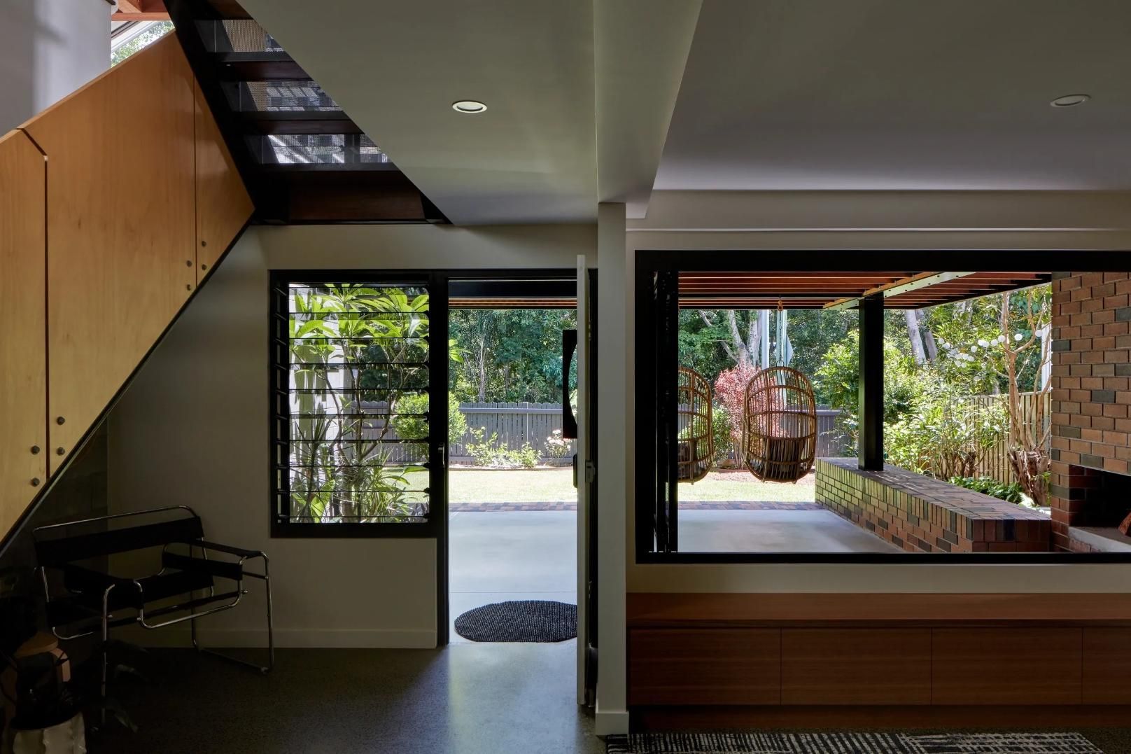 Modern living room with staircase, open doorway, and large windows overlooking a garden and patio