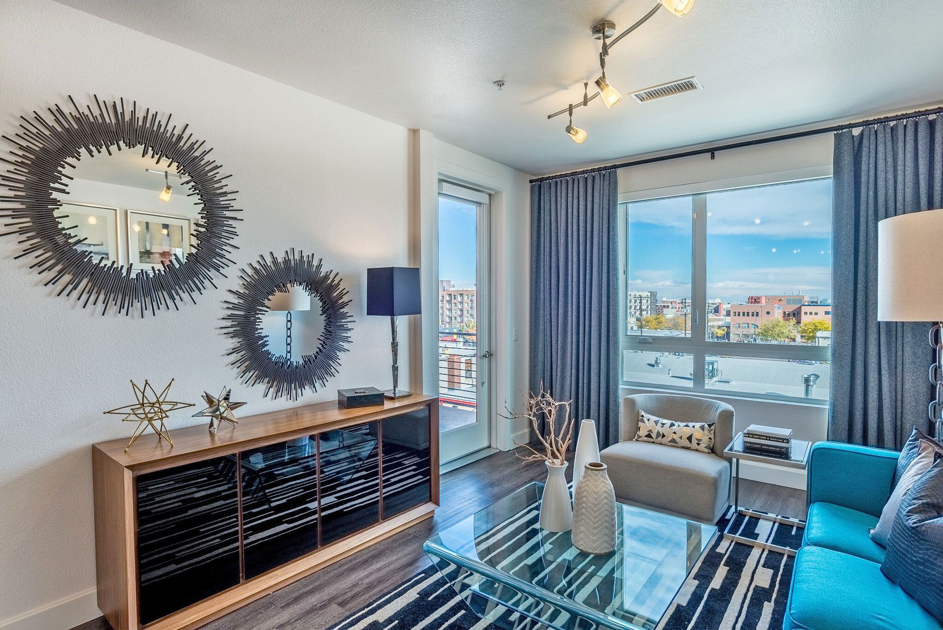 Modern living room with glass coffee table, blue sofa, and large city-view window.