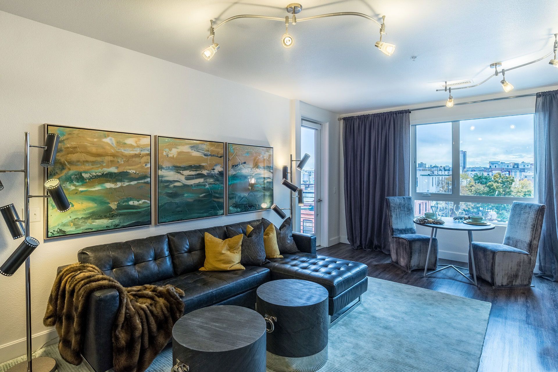 Modern living room with black leather sectional, triptych art, floor lamp, and a window-side dining nook.